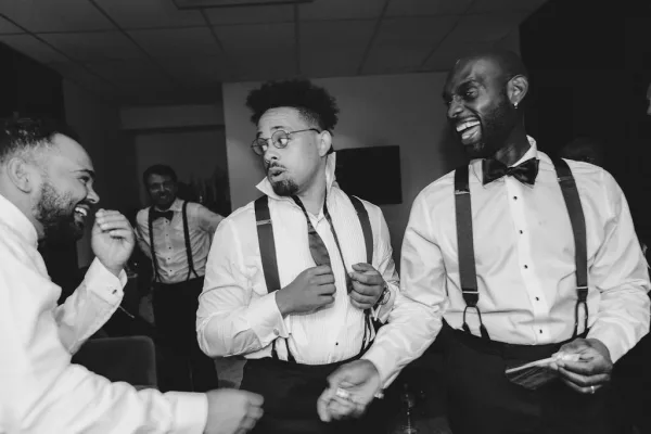 Groomsmen dancing and laughing candid in tuxedo shirts with bow ties and suspenders during an indoor reception as guests watch behind them