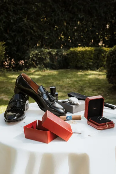 Groom accessories flat lay with black leather loafers, watch, cufflinks, tie, ring box and cigar on a white tablecloth outdoors in a garden setting