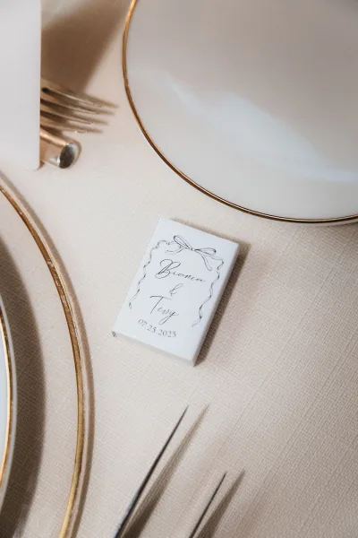 Wedding place card with calligraphy place card and ribbon illustration on a gold rim charger plate beside forks at the reception table setting