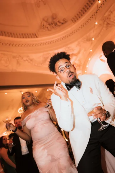 Wedding reception dancing as a man in a white tuxedo jacket and bow tie laughs with a bridesmaid holding a champagne coupe under chandeliers