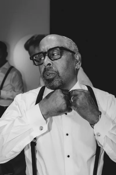 Groom getting ready adjusts bow tie over tuxedo shirt and suspenders, wearing eyeglasses and wedding band as groomsmen watch indoors