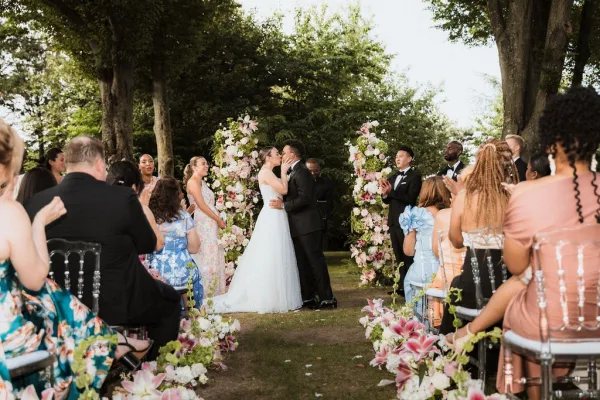 Ceremony kiss as bride in a strapless gown and groom in black tuxedo embrace under a floral arch with pink lilies in a garden setting