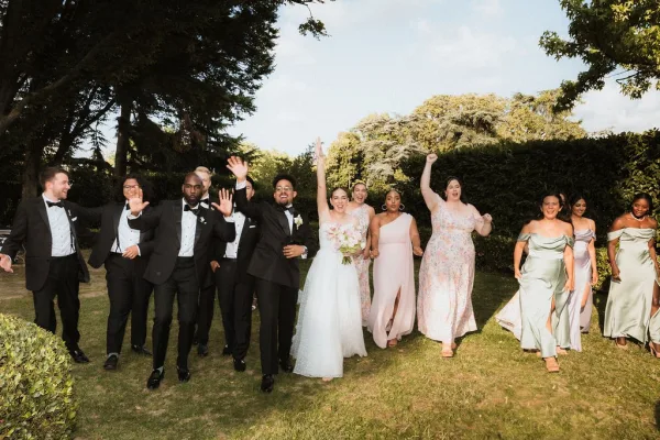 Wedding party photo of bridesmaids and groomsmen walking and cheering with the bride holding a bouquet on a lawn with hedges and trees