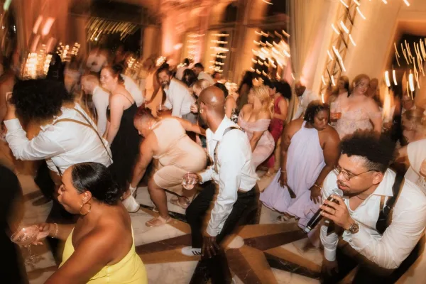 Wedding dance floor full of wedding reception dancing guests in formal attire, raising champagne glasses under warm string lights in a ballroom