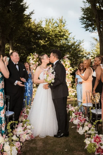 Ceremony kiss as bride in strapless gown and groom in black tuxedo embrace, bouquet in hand, with pink lilies lining a garden aisle