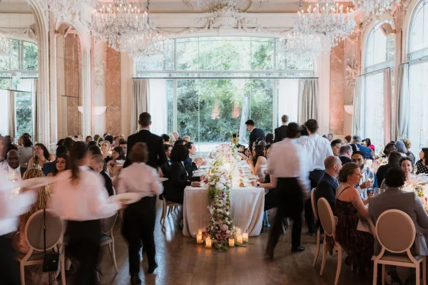 Wedding reception decor with long banquet tables dressed in white linens, floral garland, glass votives and candles beneath chandeliers in an ornate ballroom