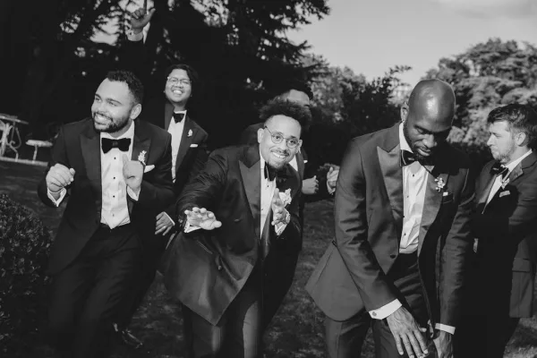 Groomsmen portrait of a groomsmen group photo in black tuxedos with bow ties and boutonnieres, posing on a lawn with trees and sky behind them