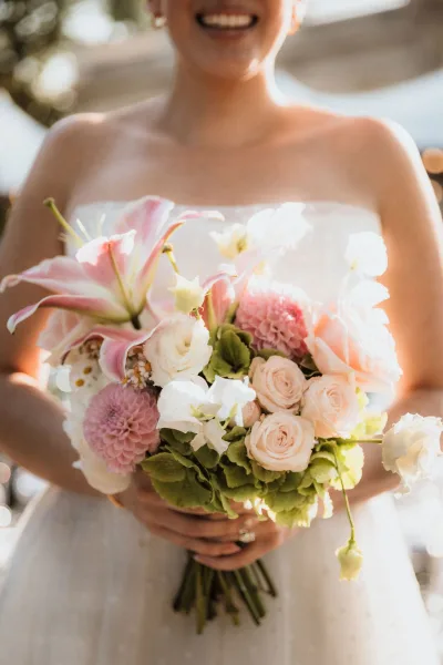 Bridal bouquet of pink lilies and pale roses with dahlias, hydrangea, and greenery held against a strapless dress with bokeh lights behind