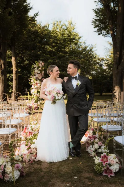 Couple portrait of bride and groom smiling arm in arm, she holds a pink lily bouquet beside a floral-lined garden aisle and arch