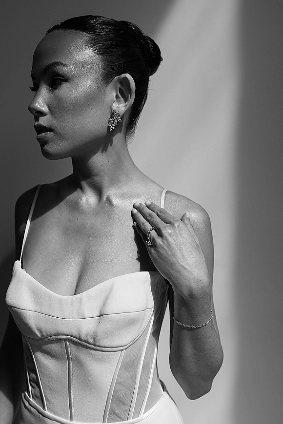 Bridal portrait in black and white showing bride in side profile with sleek updo, crystal drop earrings, corset gown by window light