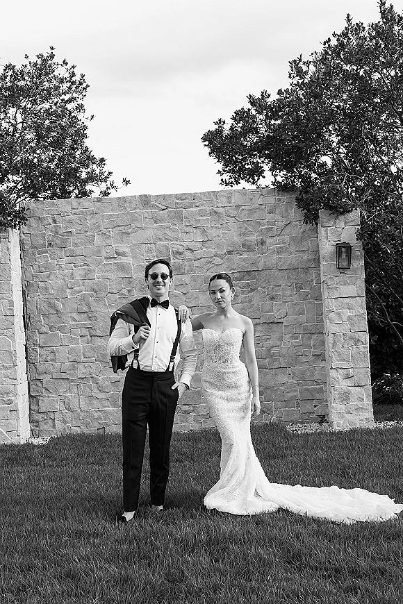 Couple portrait in a black and white wedding portrait, bride in strapless lace dress with train beside groom in tux with sunglasses by a stone wall