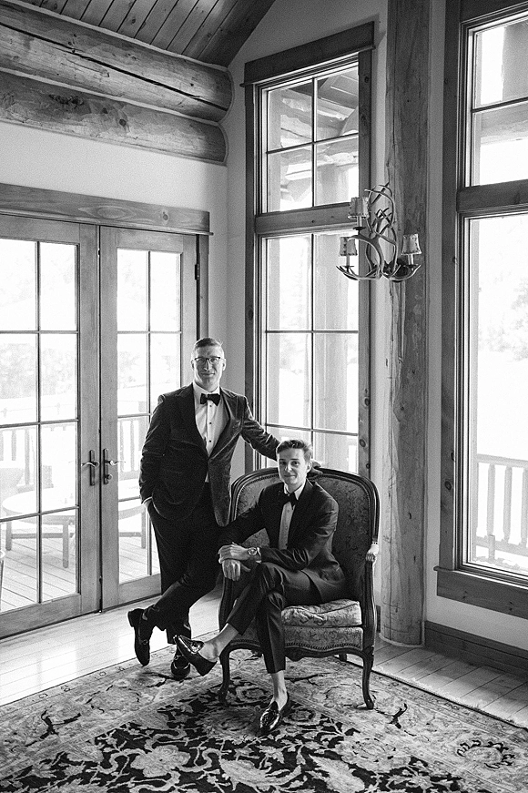 Groom portrait of two grooms in tuxedos by large windows in a rustic cabin interior, one seated in an upholstered armchair beneath an antler chandelier
