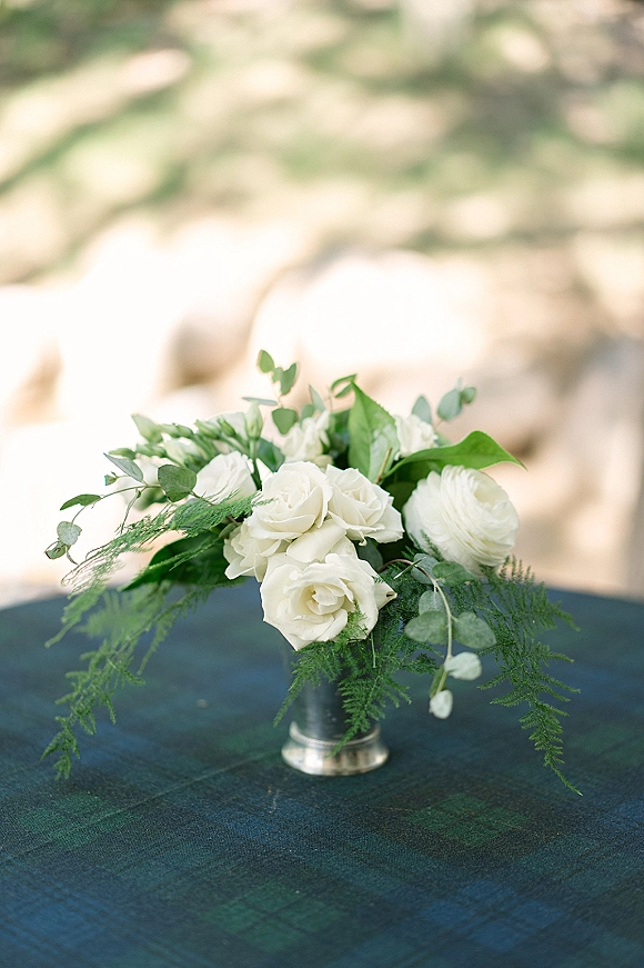 Wedding centerpiece with a white rose centerpiece of ranunculus and greenery in a silver vase on a plaid tablecloth, blurred outdoor foliage behind