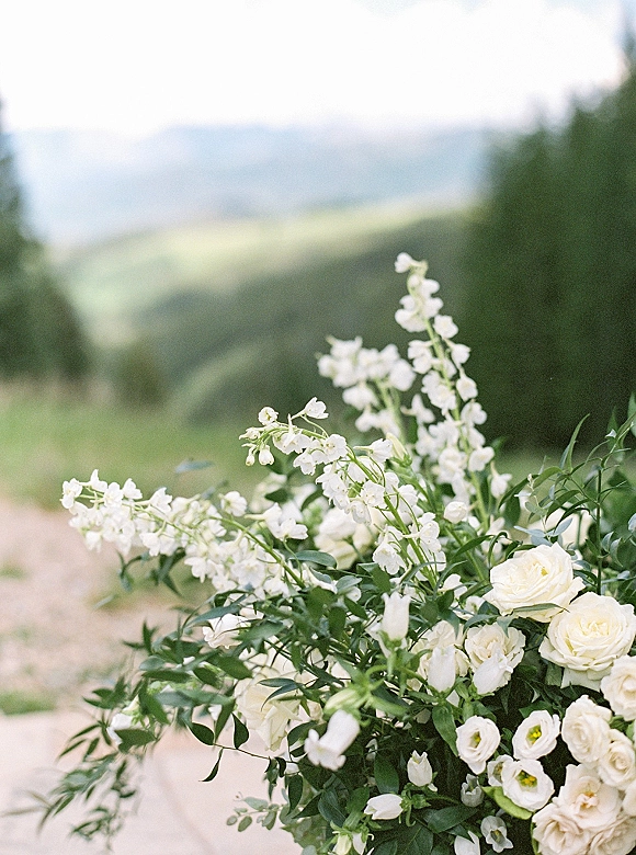 Wedding flowers in a white rose arrangement with wildflowers and greenery placed along a dirt path in a mountain meadow with evergreens