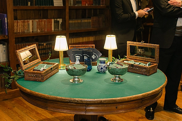Wedding cigar bar with cigars, cutters, and matchbooks on a green tablecloth round table, accented by lamps and wicker boxes in a library lounge setting