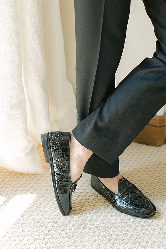Groom shoes, black wedding loafers in crocodile-embossed leather beside black suit trousers by sheer curtains in soft window light