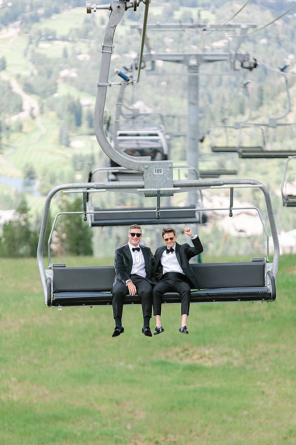Couple portrait of two grooms on chairlift in black tuxedos and sunglasses, with mountain landscape and ski lift cables behind them