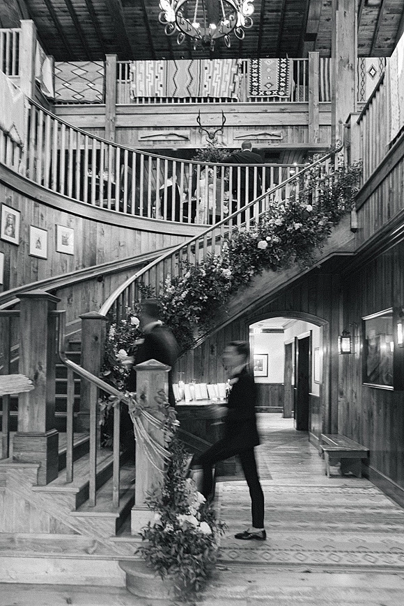Wedding staircase decor with staircase floral garland and white flowers draped on a curved railing beneath a chandelier in a wood-paneled lodge foyer