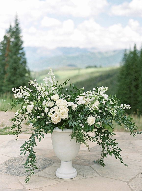 Wedding floral arrangement of white roses and greenery in a white urn vase on a stone patio with evergreen trees and mountain view