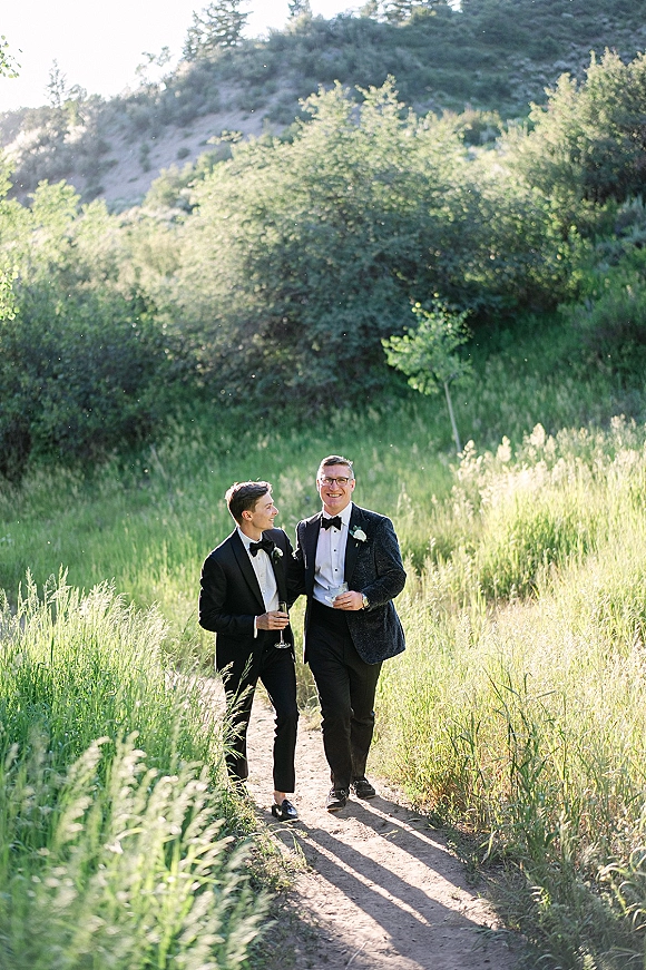 Groom portrait of two grooms in black tuxedos holding champagne flutes, walking a sunlit dirt path through tall grass and trees
