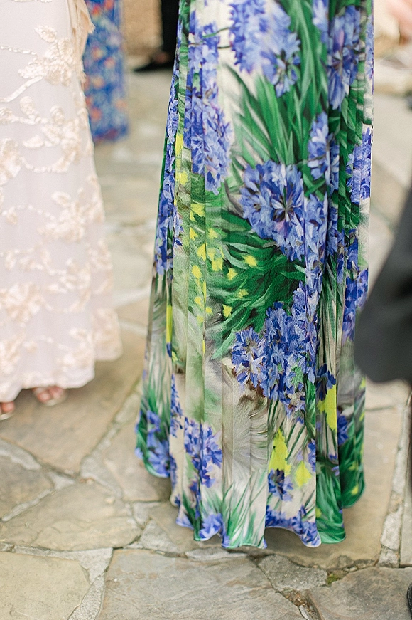 Wedding guest dress in blue floral maxi chiffon with open-toe heels, shown on a stone patio with blurred guests in the background
