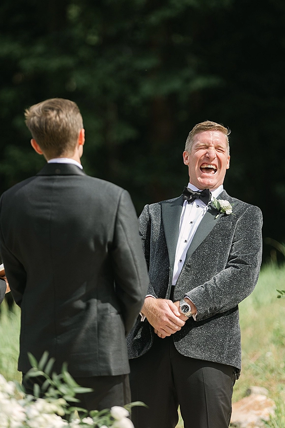 Groom reaction during a first look, smiling in a tuxedo with bow tie and boutonniere against sunlit outdoor greenery backdrop