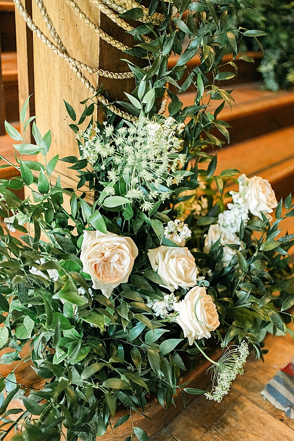 Ceremony aisle florals of white roses and greenery with a rope wrap, arranged on a wooden post beside stairs and wood flooring