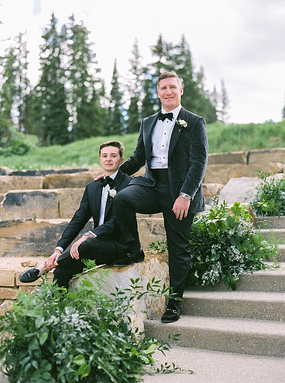 Groom portrait of two grooms in black tuxedos with bow ties and white rose boutonnieres, seated on stone steps by greenery under cloudy sky