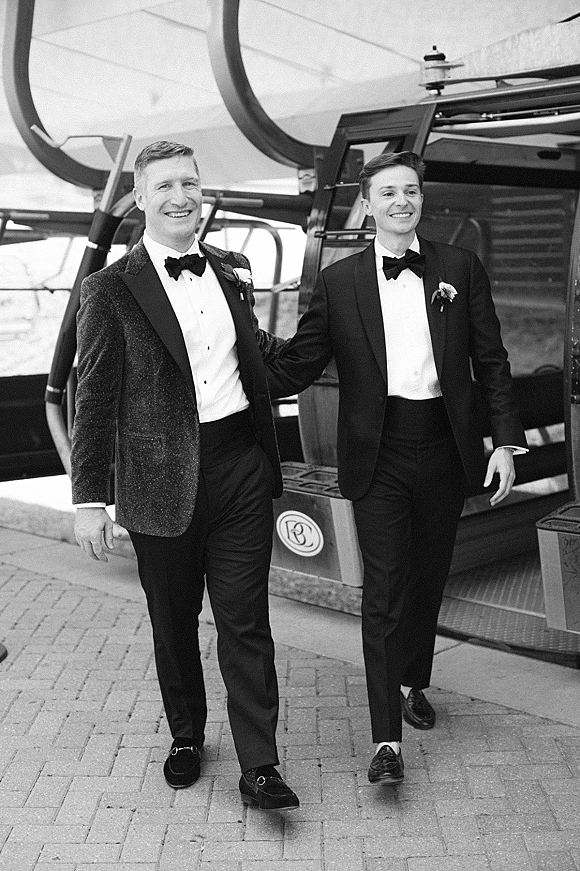 Grooms portrait of two grooms wedding photo walking hand in hand in black tuxedos with bow ties beside a gondola cabin station