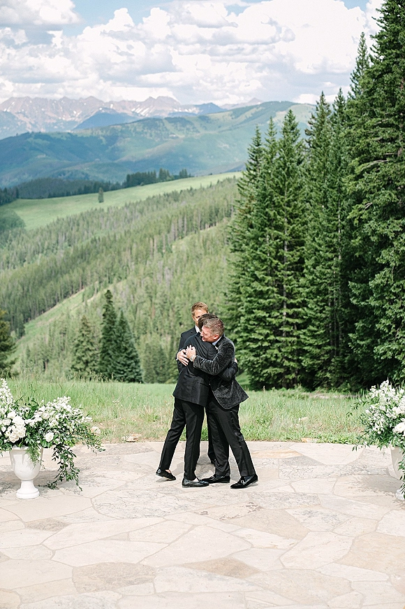 Ceremony moment as two grooms and groomsmen share a group hug in black tuxedos on a stone patio with mountain backdrop