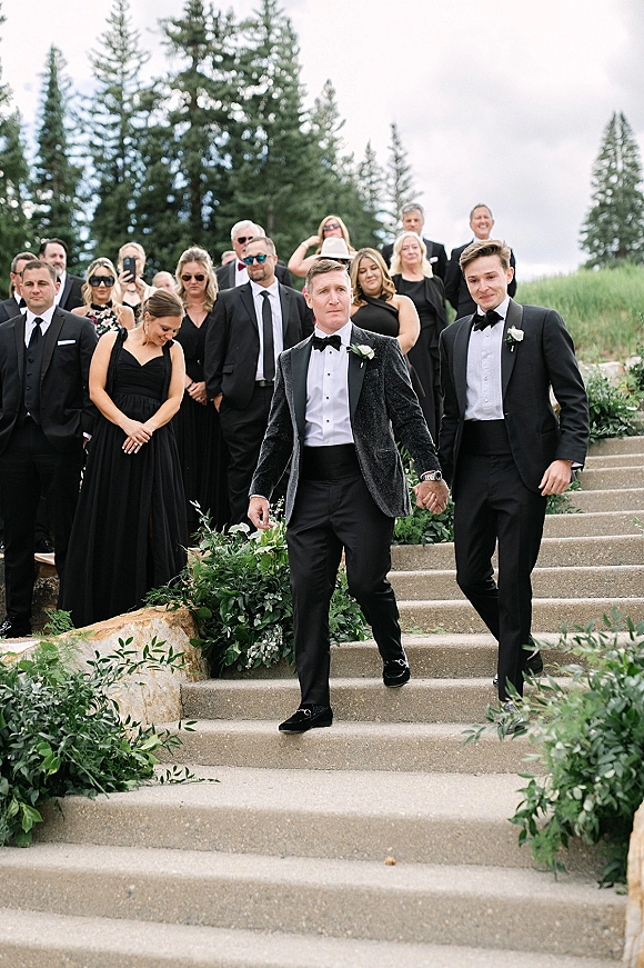 Wedding recessional with groom and groomsmen in black tuxedos and bow ties walking down stone steps by evergreens under cloudy sky