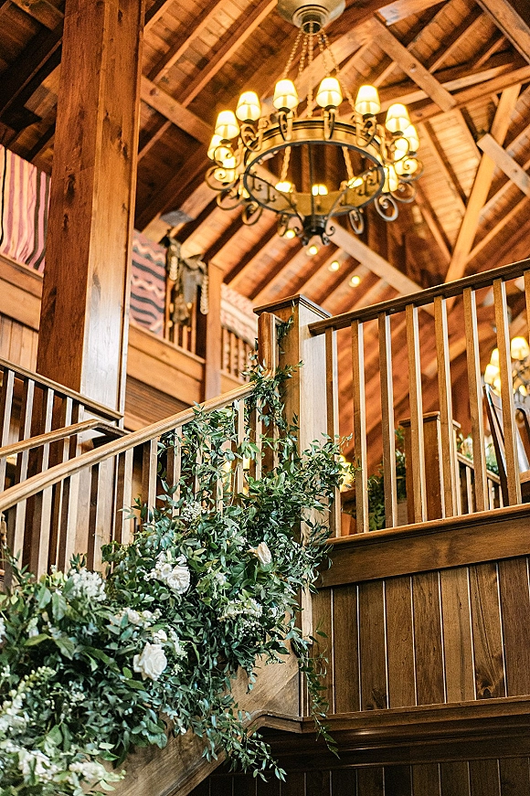 Rustic venue interior with wood beam ceiling, chandelier and string lights, plus greenery garland with white flowers on the railing