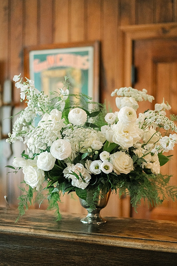 Wedding floral arrangement of white wedding centerpiece blooms and greenery in a silver urn on a wooden table against wood-paneled wall