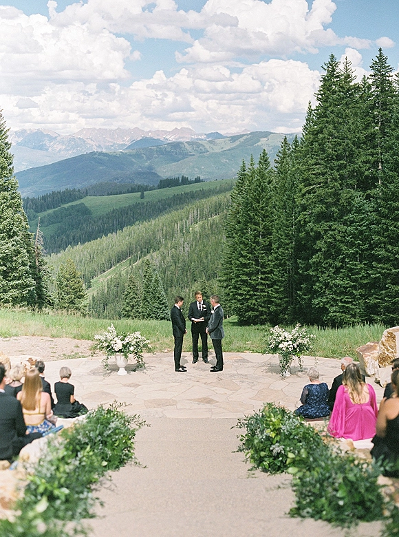 Wedding ceremony with two grooms in suits exchanging vows on a stone patio, framed by white florals and greenery, mountains behind