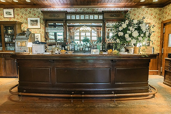 Wedding bar setup with a white floral arrangement in a silver urn, liquor bottles, glassware, and bar menu sign on a vintage wooden bar