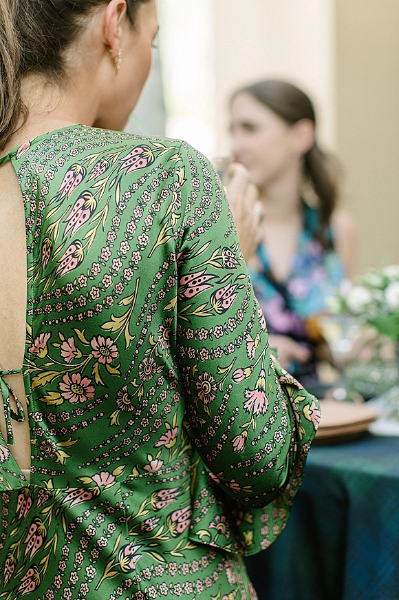 Wedding guest attire in a green floral dress with open-back tie detail and drop earrings, seated at an indoor reception table with greenery