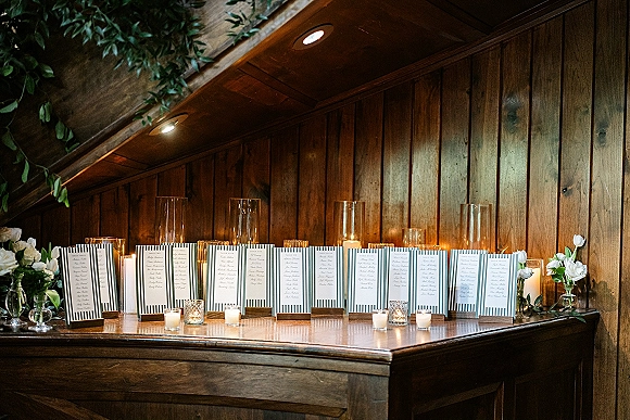 Escort card display with wedding seating chart cards in striped designs on wooden holders, accented by candlelight and greenery against a wood wall