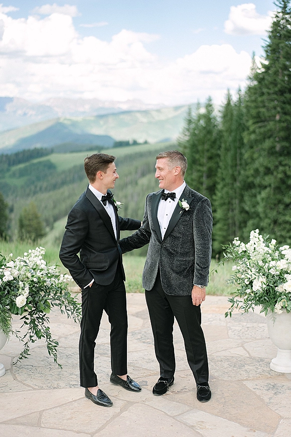 Groom portrait of two grooms in tuxedos with bow ties and white rose boutonnieres on a stone patio with mountain backdrop