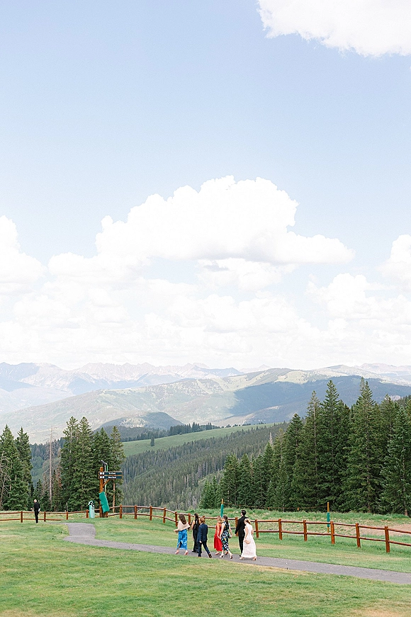 Wedding guests walking along a paved path, bridesmaid dresses adding color beside a wooden fence, with mountains and pine trees beyond
