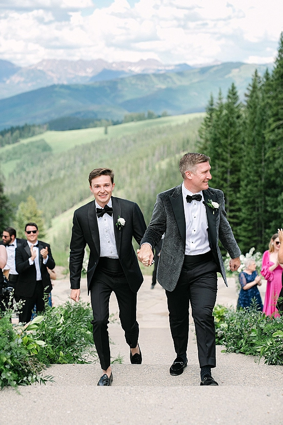 Wedding recessional with grooms holding hands in tuxedos and bow ties, walking an outdoor path as guests cheer against mountain evergreens.