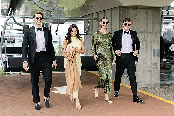 Wedding guest style with black tie wedding guests in tuxedos and dresses, walking by a ski lift station; one wears sunglasses, another a green floral dress
