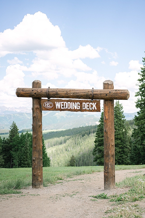 Wedding signage on a rustic wooden signboard in a log frame, hanging by metal brackets along a mountain dirt path with pines