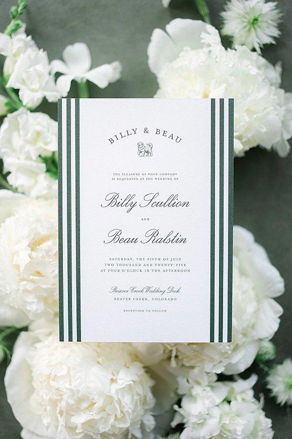Wedding invitation with classic wedding invitation styling, striped border and script calligraphy beside white peonies on a dark stone surface