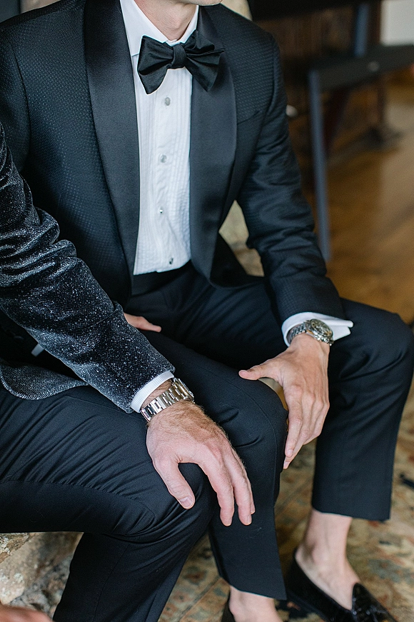 Groom tuxedo with black tie groom details, satin lapels and black bow tie, showing cufflinks, wristwatch, and dress shoes indoors