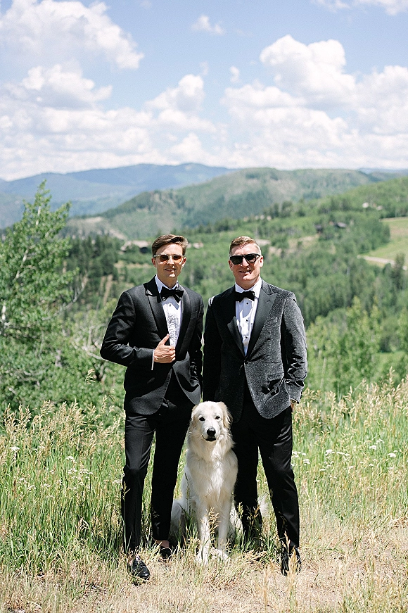 Groom portrait of two grooms in black tuxedos and sunglasses with a golden retriever, set in a mountain meadow under blue sky