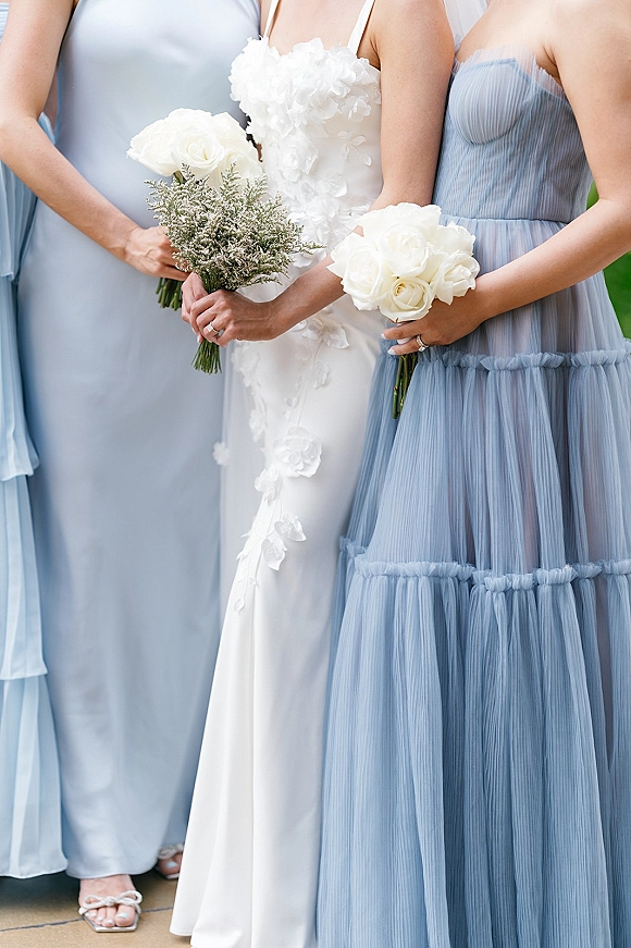 Bridesmaid bouquets of white rose bouquet blooms with greenery filler, held by women in dusty blue dresses beside a bride outdoors