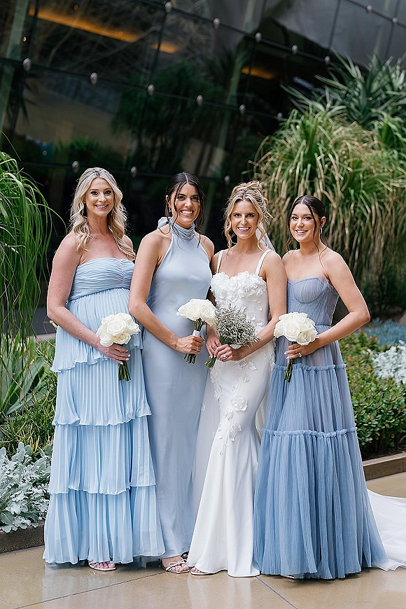 Bridesmaids portrait of the bride with bridesmaids in mismatched blue dresses holding white rose bouquets in a modern glass courtyard backdrop