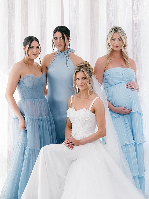 Bridesmaid group portrait with bride with bridesmaids in dusty blue dresses, bride seated in veil by sheer curtains and window light