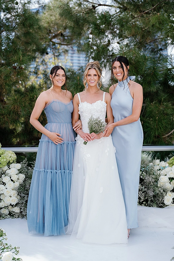 Bride with bridesmaids smiling in blue bridesmaid dresses beside a white wedding dress, holding a bouquet on a leafy outdoor railing walkway