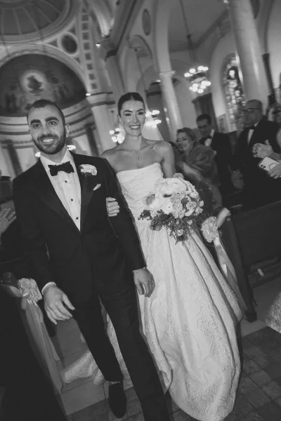 Wedding recessional with the bride and groom walking down the aisle, her bridal bouquet vivid beneath stained glass and chandeliers in a grand church.
